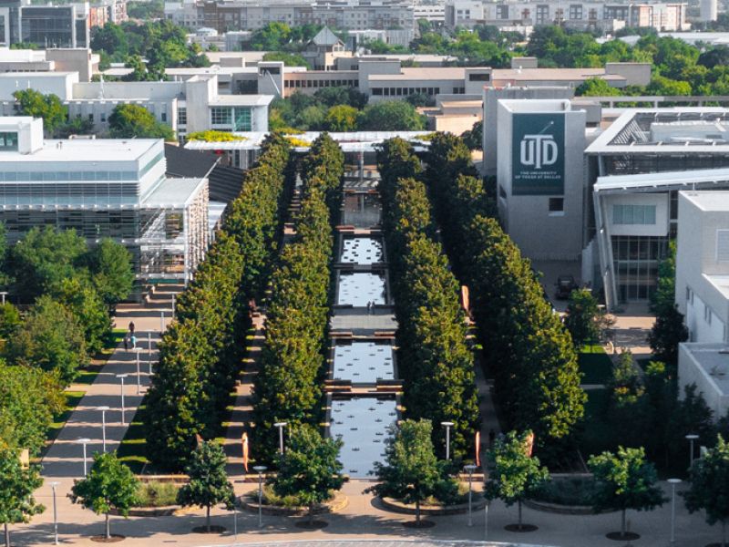 Aerial view of a campus with a tree-lined walkway and reflective pools, surrounded by modern buildings.