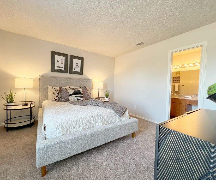 Cozy bedroom with a plush bed, side tables with lamps, and framed art. Open door to a bathroom with lit mirror.