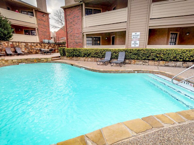 Outdoor swimming pool beside residential buildings with lounge chairs and shrubbery.