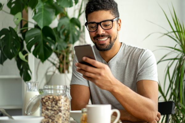 Man smiling at phone, sitting at a table with breakfast items, surrounded by houseplants.