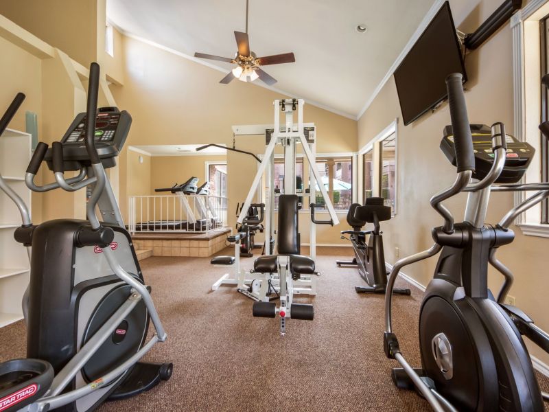 Ashwood Park gym with exercise bikes, treadmill, elliptical machines, and a weight machine, in a bright room with a ceiling fan.